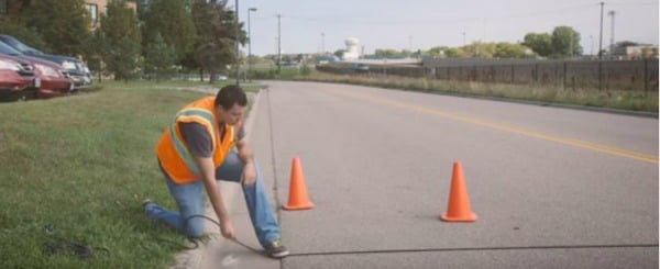 Field worker kneeling at a curb installing a pneumatic tube counter on the roadway with two traffic cones in the background